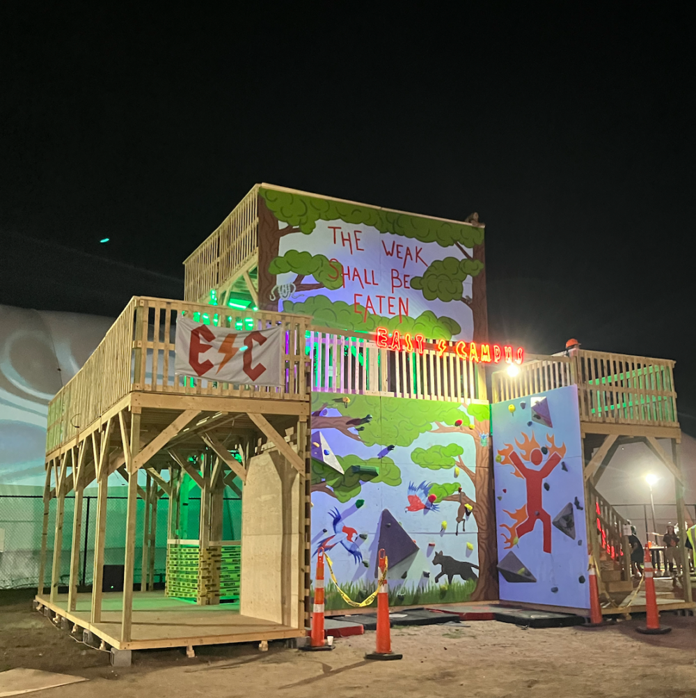 A colorful, painted with murals, lit-up three-story wooden fort and rock climbing wall. There's an LED sign that says EAST CAMPUS, a banner that has E C with a lightning bolt, and the motto "the weak shall be eaten" on top.