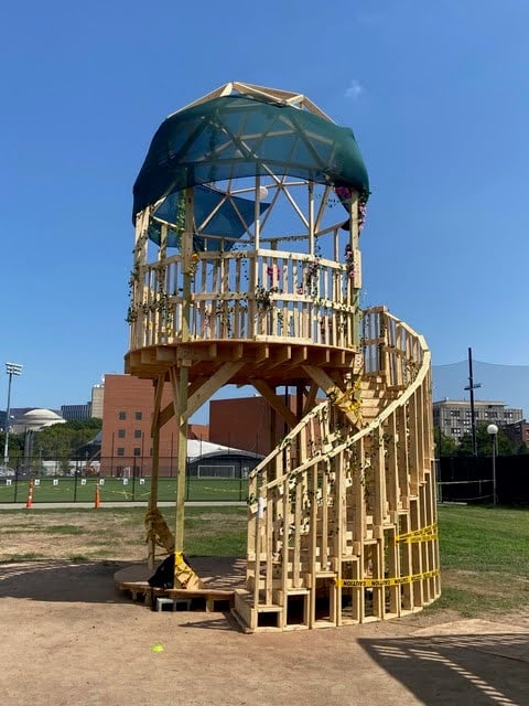 A wooden treehouse with spiral stairs and topped with a fabric geodesic dome.