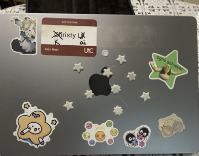 A macbook with a lot of stickers on it. From left to right: Top row: A sticker of Barbara from Genshin Impact; a "Meet your UAC Advisor" Nametag. Middle row: A bunch of puffy star stickers; a sticker of Shrek in a green star with the phrase "ogre achiever" in comic sans. Bottom row: A cute character wearing a star around his head; seven smily faces and the word "smile"; two dust mites and some shapes in blue, purple, and magenta; a faded sticker of three spherical animals. 