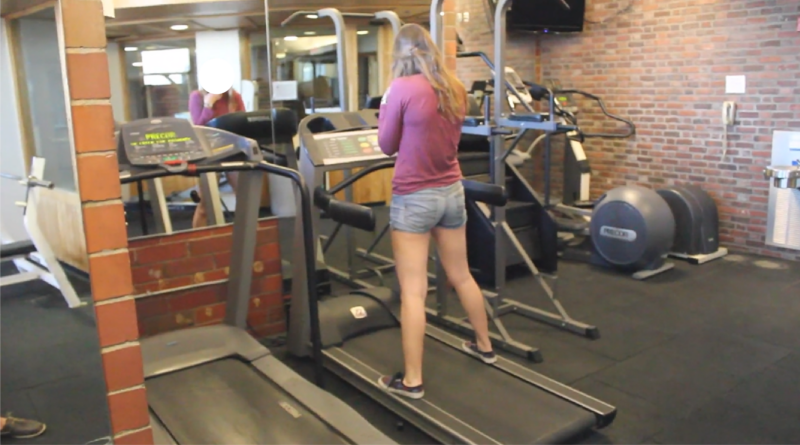 a picture of a lady standing on a treadmill in a gym with a brick wall