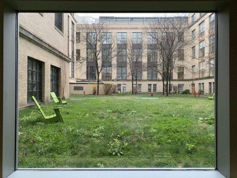 an area of mit surrounded by beige walls. inside is a grass field with a few green lawn chairs and trees without leaves.