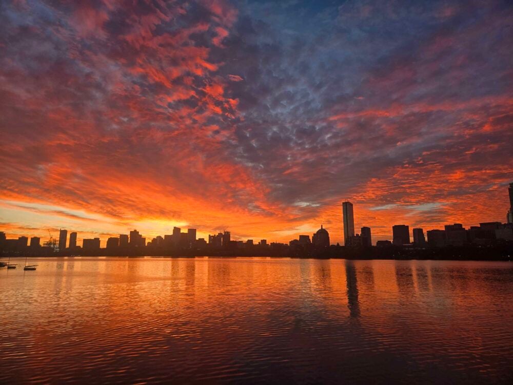 a bright orange and pink sunrise over a dark skyline and reflected in the charles river