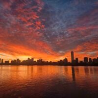 a bright orange and pink sunrise over a dark skyline and reflected in the charles…