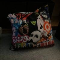 A decorative pillow featuring logos of popular musicals like Wicked, Phantom of the Opera, and…