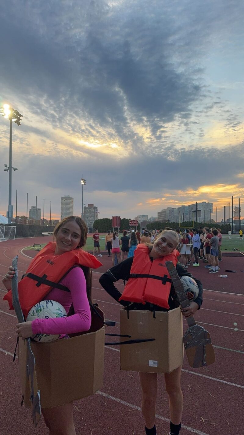Two girls dressed up as canoers smiling next to a sunset.