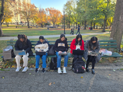 five people sitting at a bench in a park typing furiously