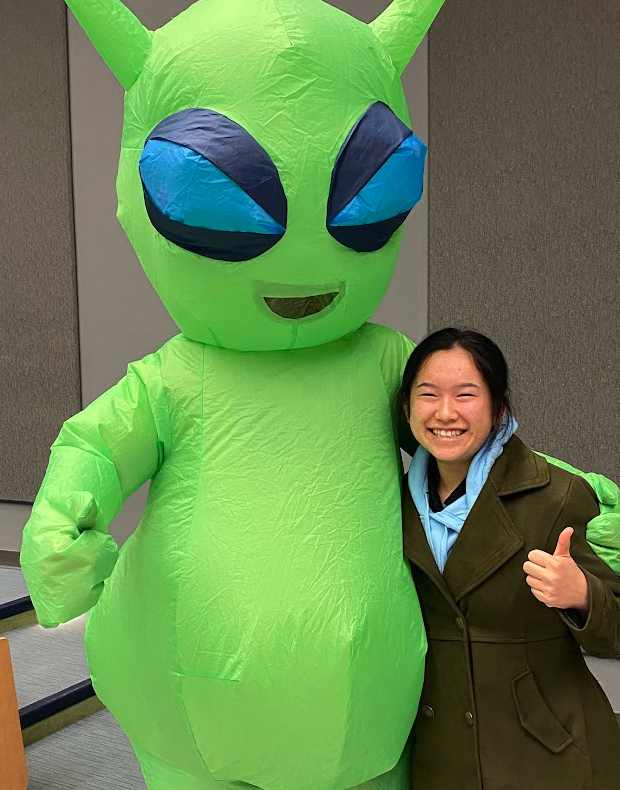 person standing next to other person in green inflatable alien costume. both people holding thumbs up