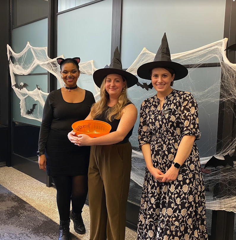 three people standing in witch and cat costumes, person in middle holding a box of candy