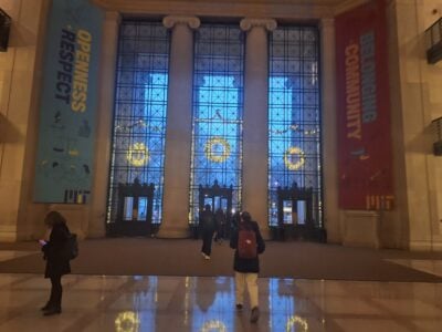 A photo of the inside of lobby 7 decorated with three lit wreaths on the entrance windows between the pillars.