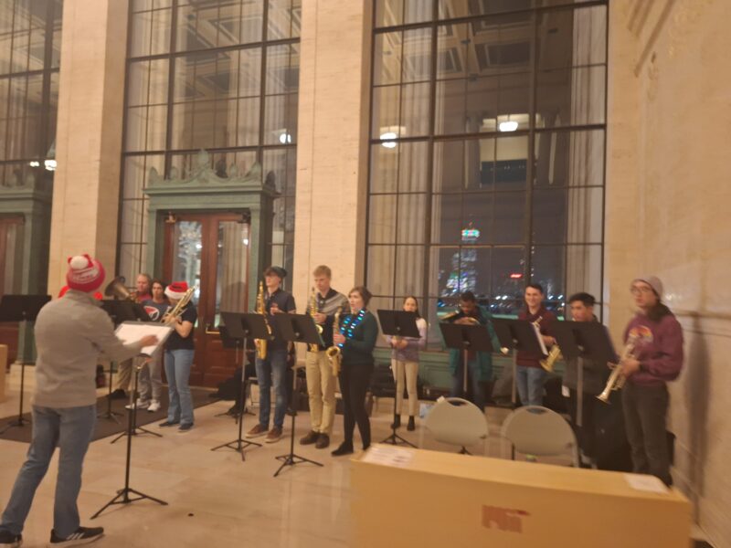 A standing orchestra playing Christmas music in lobby 10. The musicians are in a circle by the windows that overlook Killian.