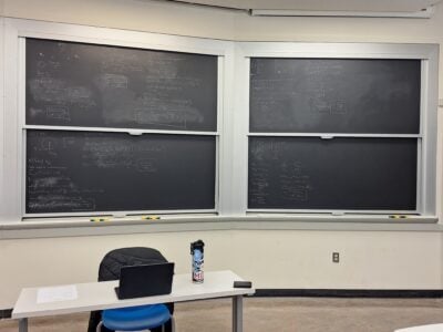 A classroom showing four chalkboards covering in practice problems for mechanical physics and a desk with a student's belongings.