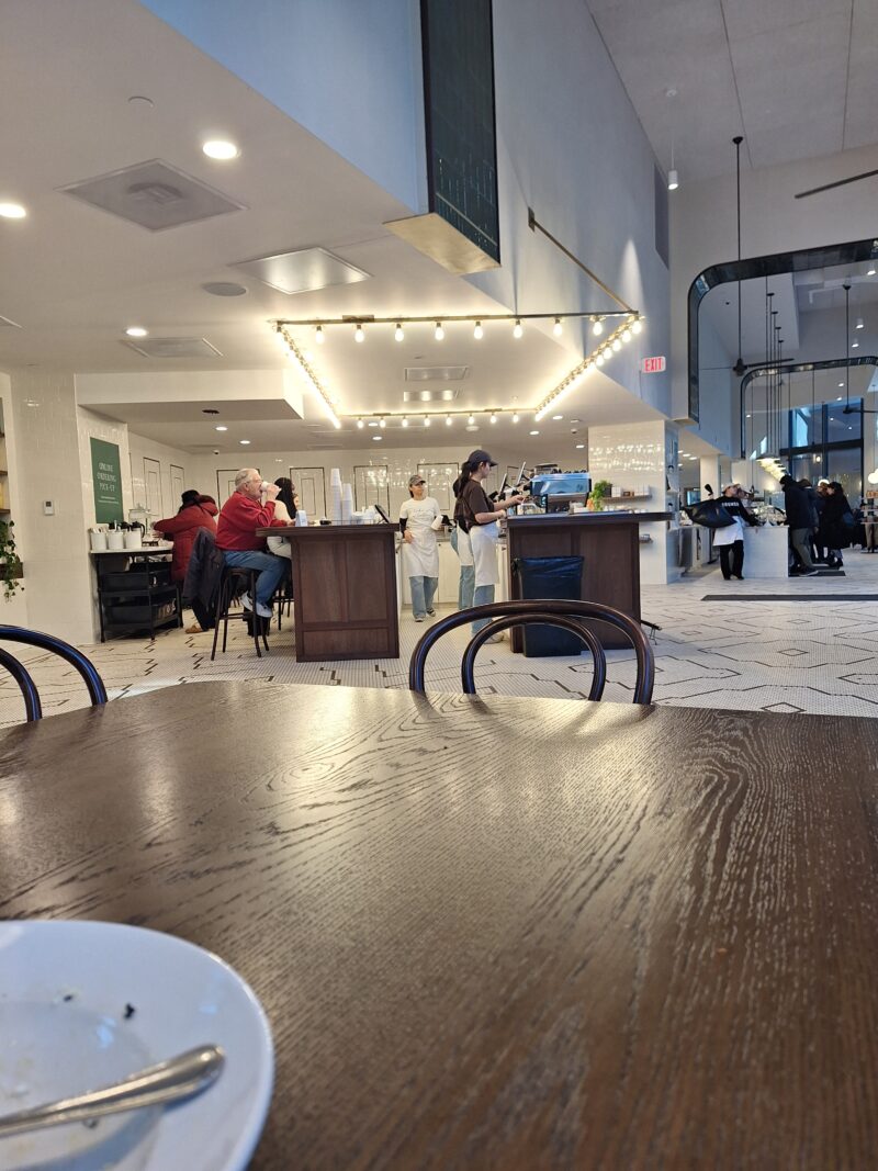 The view of a cafe from someone sitting at a long wooden table. In the middle, baristas prepare coffee at the bar.