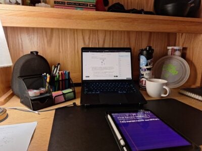 A student's wooden desk with an iPad and MacBook open to physics problems. The desk contains office supplies, a lamp, books, and a frisbee.