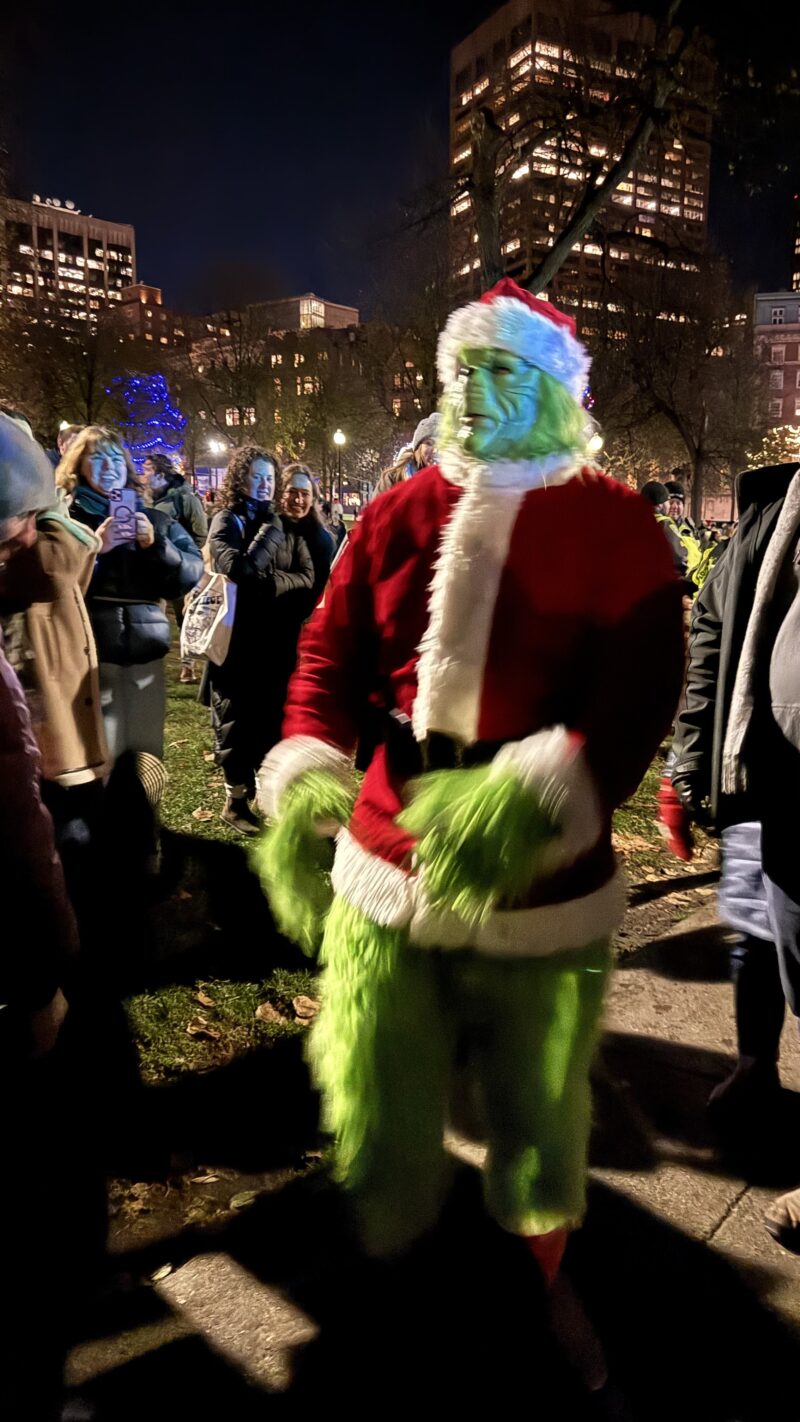 A grinch impersonator wearing a Santa outfit on the sidewalk.