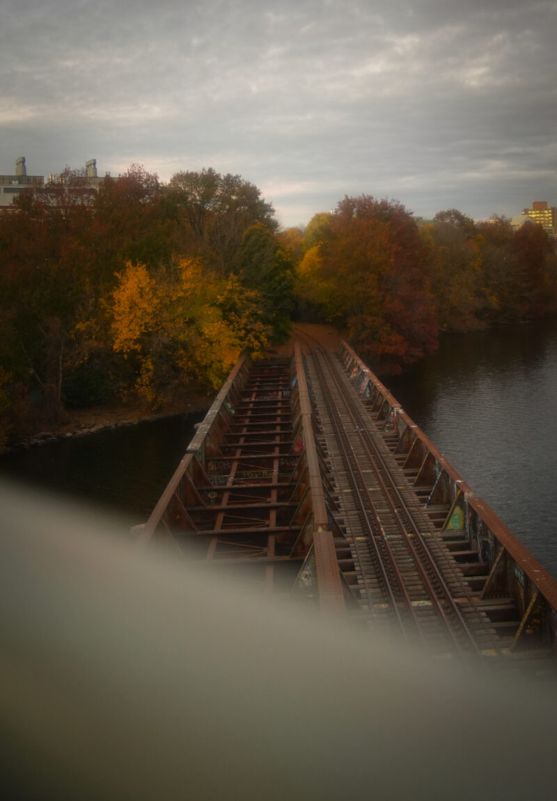 Photo of train tracks over the Charles river in front of trees during the fall