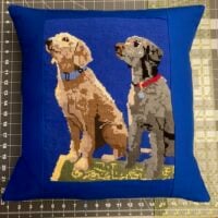 Pillow with needlepoint of two dogs on a blue background, placed on a cutting mat.