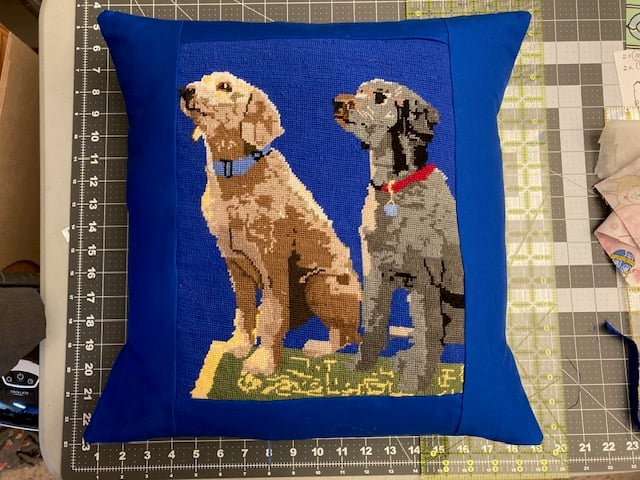 Pillow with needlepoint of two dogs on a blue background, placed on a cutting mat.