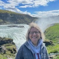 Smiling woman in glasses and a gray jacket standing by a waterfall with greenery and…