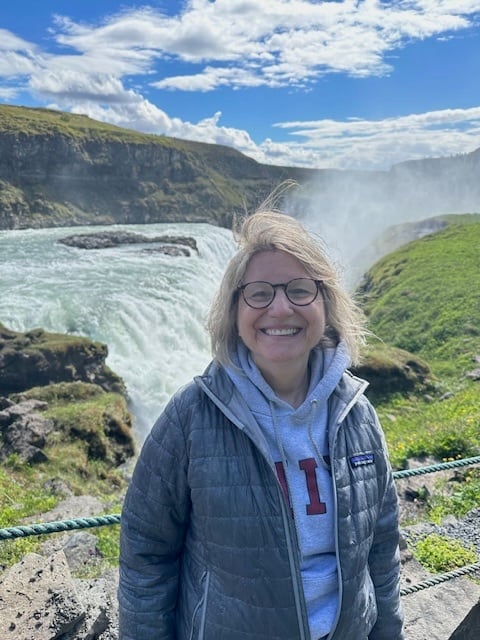 Smiling woman in glasses and a gray jacket standing by a waterfall with greenery and a blue sky.