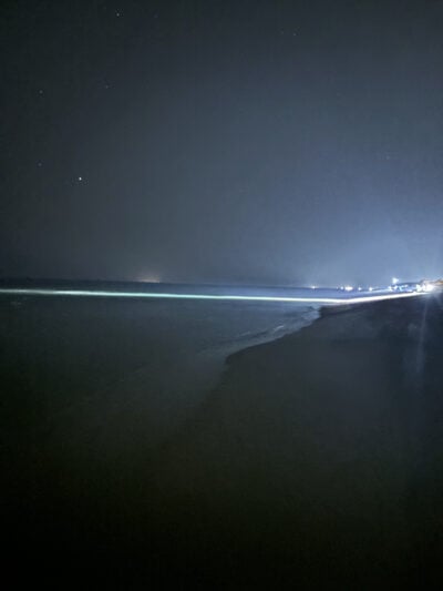 A long exposure photograph capturing a nighttime beach scene. The foreground shows a dark sandy beach that gently curves into the ocean. The waves are faintly lit, reflecting light from the horizon. The background features a dimly lit shoreline with distant lights, while the sky is predominantly dark with scattered stars, creating a moody and tranquil composition. The overall feel is calm and mysterious, emphasizing the contrast between the dark beach and the illuminated water.