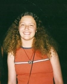 Teenager with chest-length curly bronze hair grinning and wearing an orange tank top