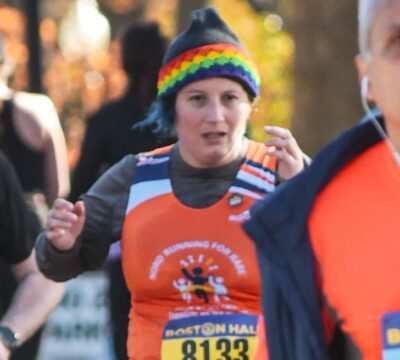 A person in an orange singlet over a brown long-sleeved shirt running in a race. They have blue hair poking out from under a gray beanie with rainbow trim. There are woods in the background.