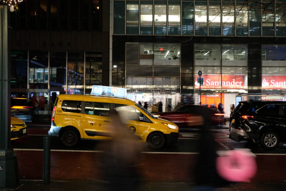 a street at night, blurred pedestrians in front of a tax in front of other blurred cars