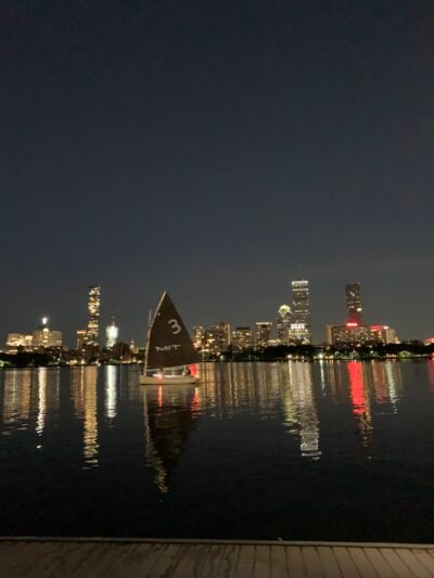 a river at night with a sailboat and reflections of tall buildings on the other side 