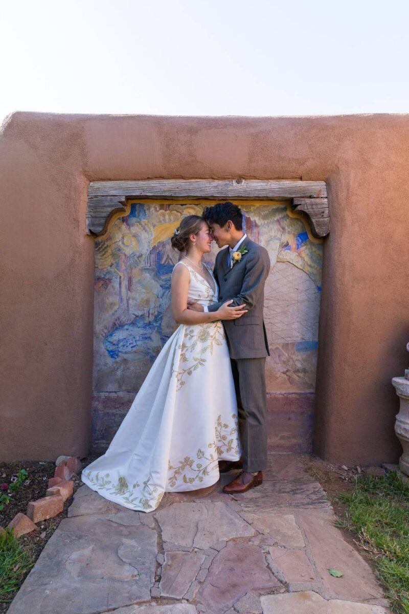 A woman and a man embracing in front of a mural inset on an adobe wall. The woman (the author) is wearing a long voluminous white wedding dress with gold embroidery and the man is wearing a gray and brown suit.