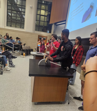 One team member presenting into a microphone and another demonstrating a device to the audience.