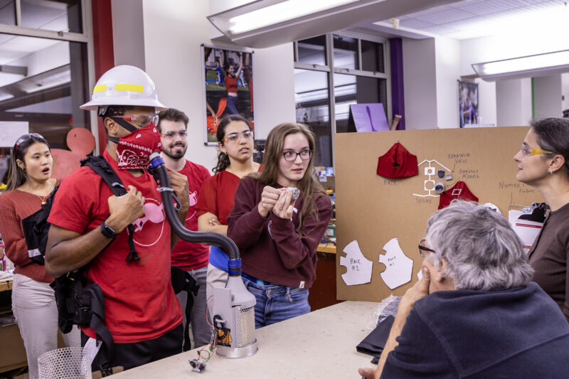 A person in a hard hat and red shirt demonstrates a device with a tube while others watch in a workshop.
