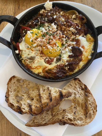 A skillet of shakshuka with two sunny-side-up eggs, crumbled cheese, and herbs, served alongside two slices of toasted rustic bread on a white plate.