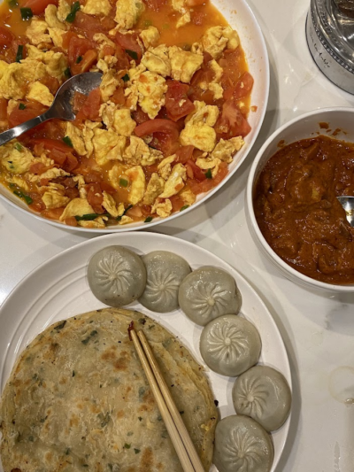 A meal spread featuring tomato and scrambled eggs in a bowl, steamed dumplings, a flat scallion pancake with chopsticks, and a small bowl of curry on a white table.