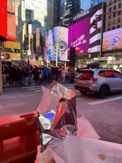 A foil-wrapped street food item held up in Times Square, with bright billboards, a crowd of people, and cars in the background.