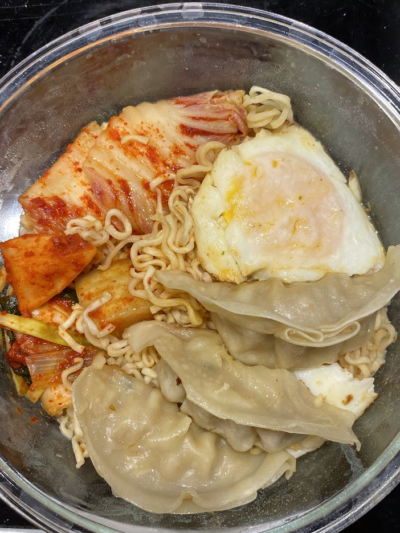 A bowl of instant noodles topped with kimchi, a fried egg, and several steamed dumplings.