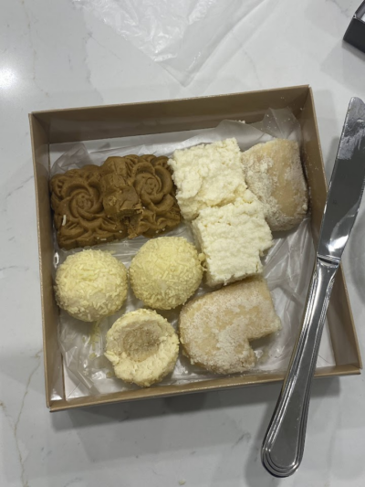 A box of assorted pastries, including mooncakes, coconut-covered balls, and powdered desserts, placed on a white marble countertop with a knife beside it.