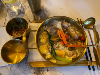 A golden pot of seafood hotpot featuring prawns, vegetables, and noodles in broth, accompanied by dipping sauces and golden utensils on a wooden tray.