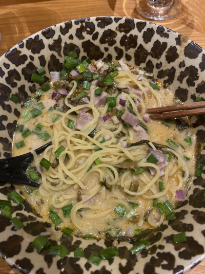 A bowl of creamy noodle soup topped with chopped green onions, diced red onions, and slices of ham, served in a patterned bowl with chopsticks and a spoon.