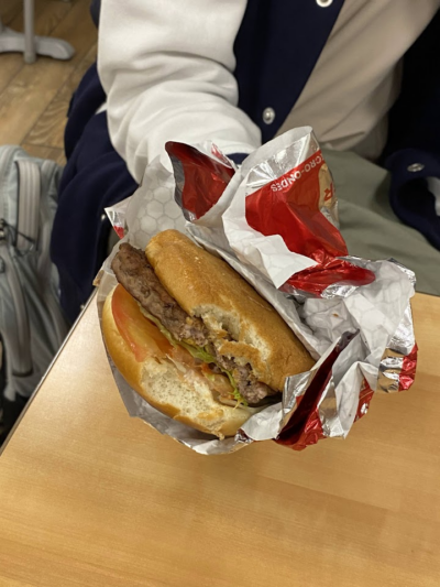 A partially unwrapped hamburger with a beef patty, lettuce, and tomato, held by a person wearing a navy and white jacket, placed on a light wooden table.