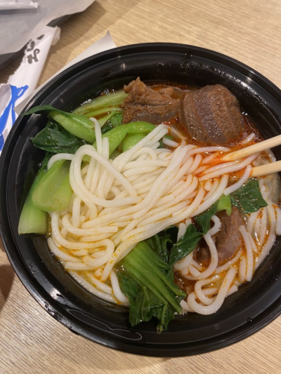 A bowl of beef noodle soup featuring thick noodles, bok choy, chunks of tender beef, and a red broth, served in a black disposable container.