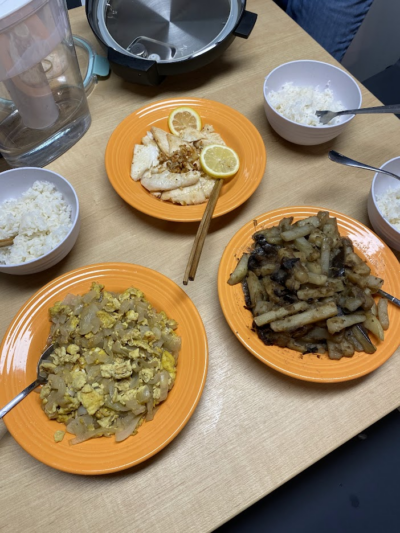 A table setting with three orange plates containing stir-fried dishes, including scrambled eggs with onions, sautéed vegetables, and pan-fried fish garnished with lemon slices, accompanied by bowls of white rice.