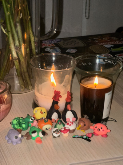 A collection of small, colorful clay figurines, including animals and mushrooms, placed in front of two lit candles in glass holders on a table.