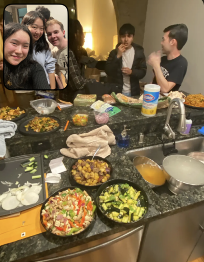 A lively kitchen scene with a group of people chatting and eating around a counter filled with various dishes, including salads, roasted vegetables, and snacks, with an inset selfie of smiling friends in the top left corner.