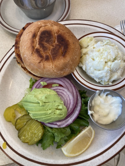 A plate featuring a toasted burger bun, avocado slices, red onion, lettuce, pickles, and a side of potato salad, served with a lemon wedge and a small cup of sauce.