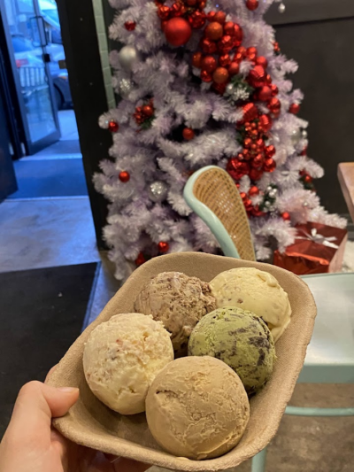 A bowl containing five scoops of assorted ice cream flavors, held in front of a white Christmas tree decorated with red and silver ornaments.