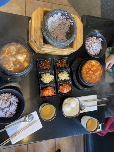 A Korean meal spread featuring stone pots of purple rice, bubbling stews, kimchi, and side dishes like beans and sprouts, served on a black tabletop.