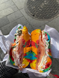 A colorful rainbow bagel sliced in half, filled with cream cheese, smoked salmon, and capers, held over a city sidewalk.