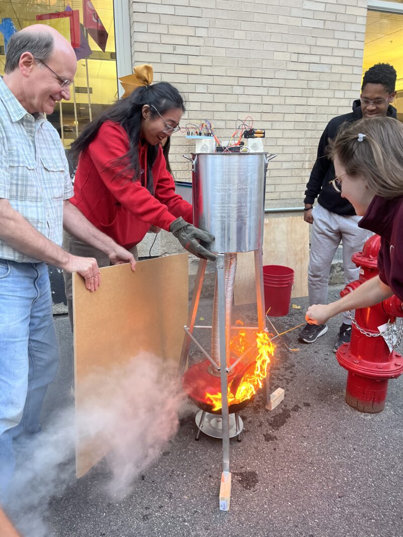 People surrounding a metal test rig with a fire at the bottom.