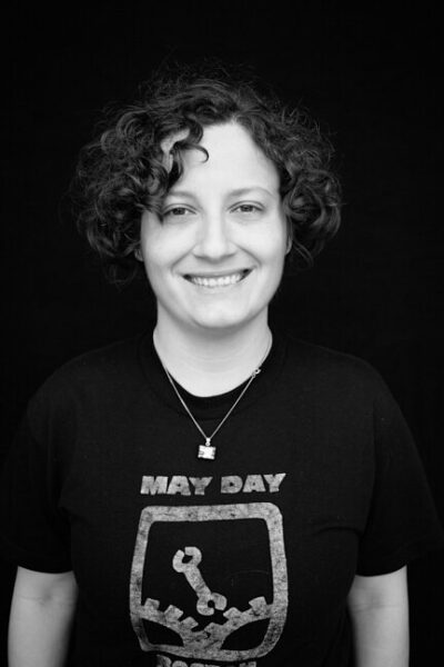 Black-and-white photo of a person with curly dark ear-length hair in a "May Day 2012" t-shirt