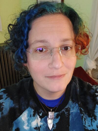 A person with blue, green, and orange curly ear-length hair, blue-light-reflecting glasses, and a blue/black tie-dyed sweatshirt